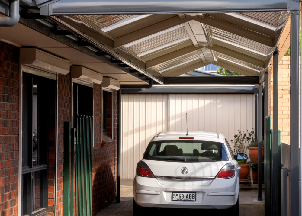 White car parked under a gable-style metal and polycarbonate carport attached to a brick house with paved driveway.