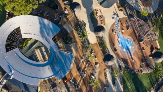 Aerial view of a modern architecture project featuring a curved steel roof, landscaped pathways, greenery, and a nature-inspired playground.