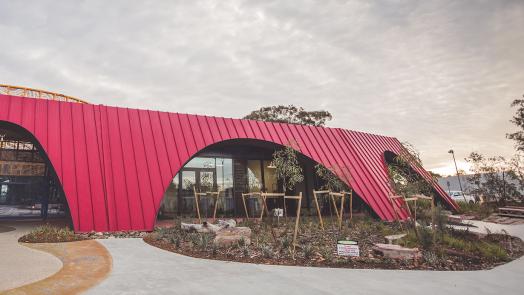 A modern building featuring a bold red facade made of KLIP-LOK 700 HI-STRENGTH® metal sheeting, with distinctive curved archways. Large glass windows and doors reflect the surrounding trees and landscape.